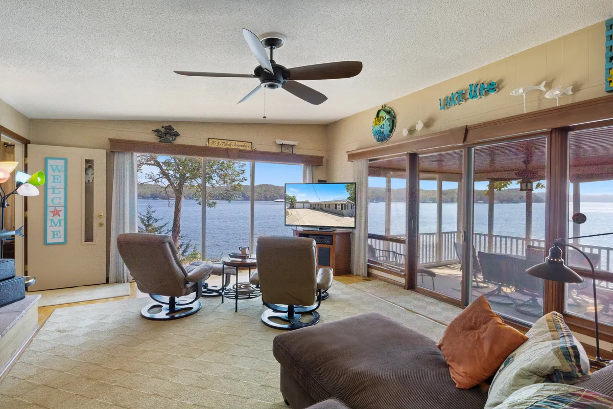 This inviting living room features large windows that flood the space with natural light and offer scenic lake views. A comfortable seating area is arranged around a television, blending relaxation and entertainment with the beauty of the outdoors.