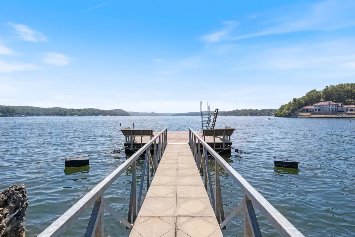 This image features the private dock extending over the water, with visible boat slip, swimming access, and ample space for fishing or relaxing. Houses in the background add a sense of peaceful lakeside community, and the sunset light across the water enhances the inviting and serene mood.