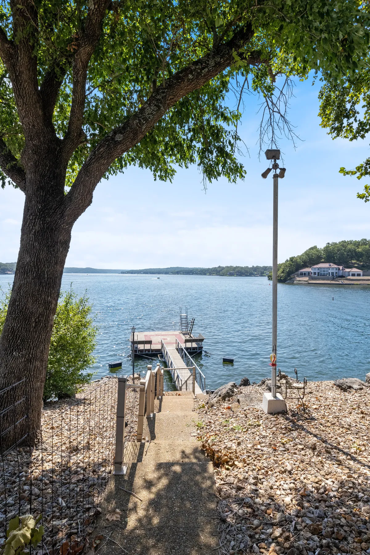 A serene shot of a large tree shading a sturdy dock that stretches over peaceful water. The gentle path leading to the dock is inviting, suggesting easy access for fishing, boating, or simply enjoying the natural lakeside environment.