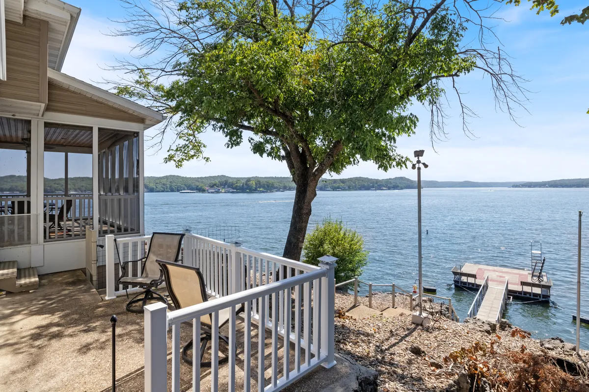 A tranquil lakeside scene featuring a boat dock framed by a large tree, comfortable deck furniture, and expansive water views. The setting is ideal for activities like fishing, boating, and relaxing by the water with friends or family. Subtle lighting hints at the magic of both sunrise and sunset gatherings.