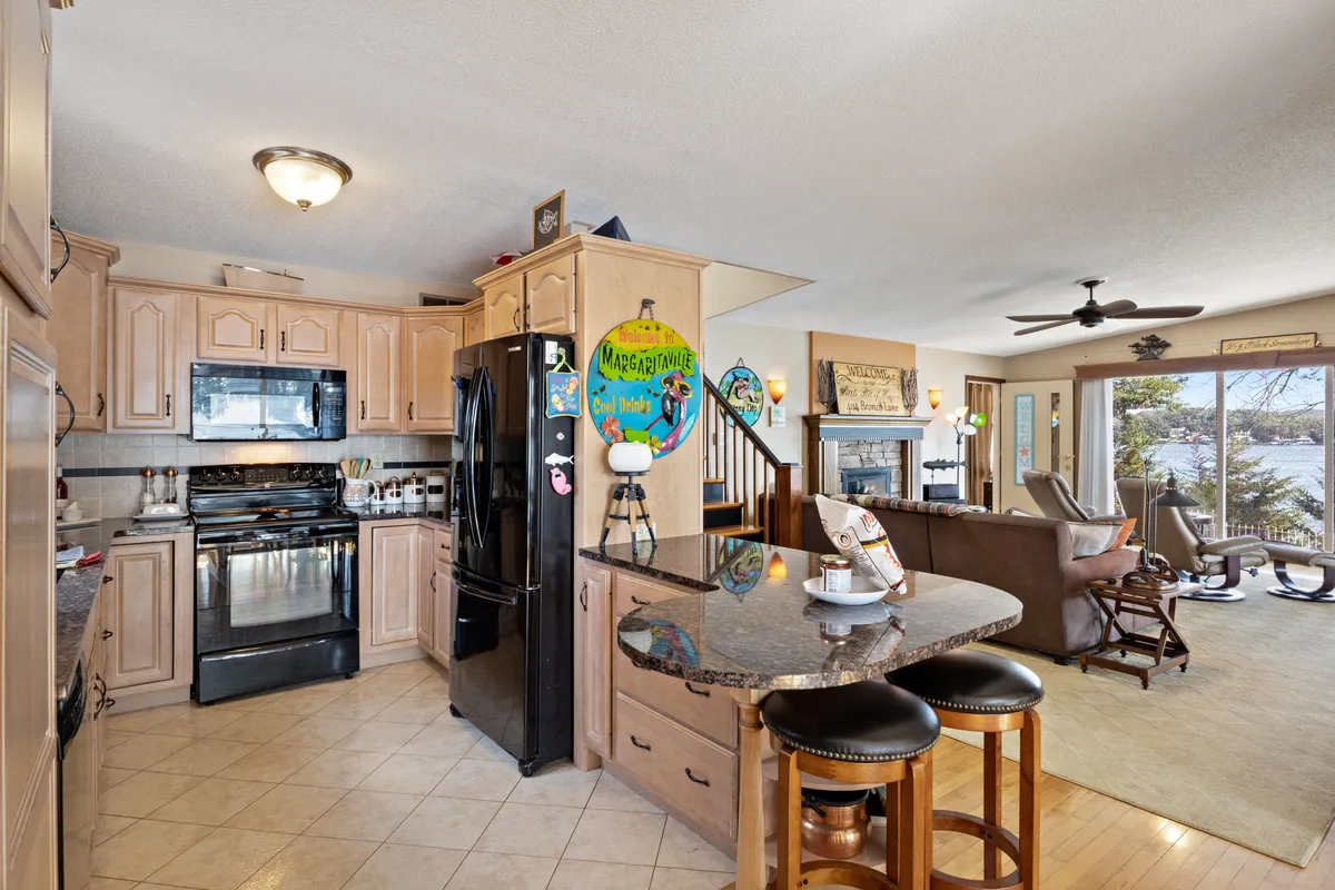 An open-plan kitchen with a spacious central island, black appliances, barstools for casual seating, and windows framing the water beyond. The kitchen seamlessly connects with the adjacent dining area, inviting both cooking and conversation against a backdrop of nature.