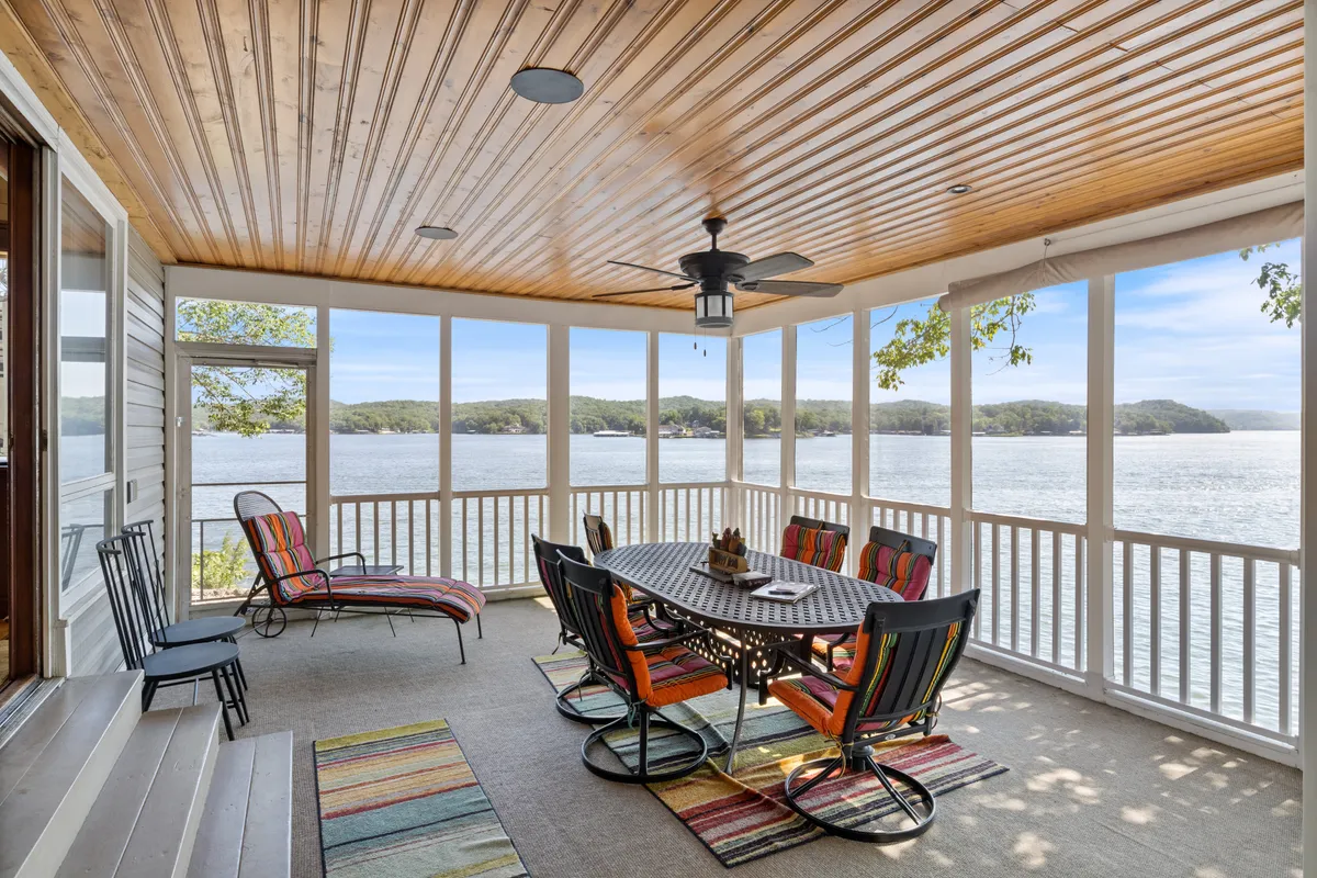 The image features a spacious outdoor dining area set with a large table surrounded by colorful chairs, overlooking a serene lake. The view includes sweeping sunset hues reflected on the water, with lush greenery and an inviting atmosphere ideal for social gatherings. The tranquil setting suggests space for both relaxation and lively outdoor meals, capturing the essence of lakeside living.
