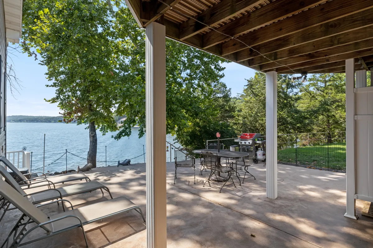 A casual lakeside patio with ample outdoor seating, a grill area, and a large tree providing shade. The scene overlooks the water and conveys a sense of group enjoyment, ideal for outdoor dining, grilling, or sunbathing with loved ones during summer evenings.